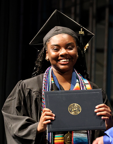 A UIS graduate smiles while holding her diploma on stage during the commencement ceremony, wearing a cap and gown.