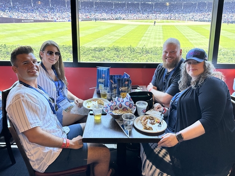 four people sit at a table with a baseball field outside the window behind them