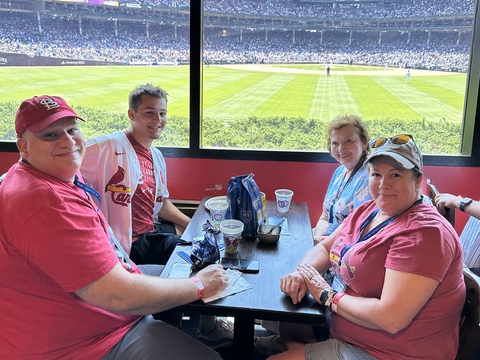 four people sit at a table with a baseball field outside the window behind them