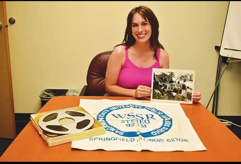 Sangamon Experience Graduate Assistant Evie holds a photo, sitting with a printed WSSR cloth and audio reel on a table.