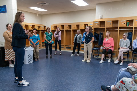A group of people gathered in a locker room, listening to a speaker.