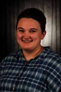 Morell Old, smiling in a blue plaid shirt against a dark background.