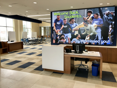 A campus lounge with a large digital welcome screen and a front desk; additional details described in the text below.
