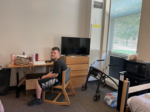 A man sitting at a desk with items scattered around the dorm room.