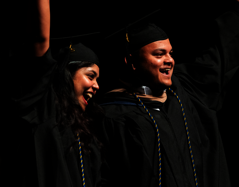 A male and female waving to the crowd in their graduation gowns.