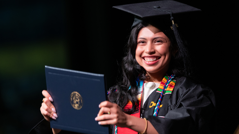 Graduating person smiling, holding diploma, wearing cap and gown.
