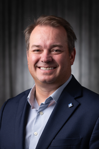 Rusty Jones smiling in a dark suit and blue shirt against a dark background.