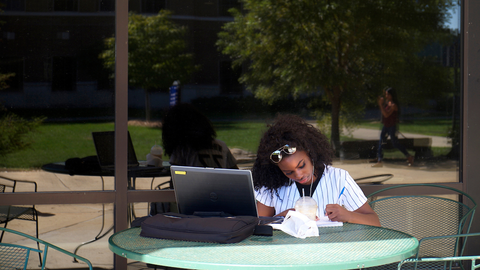 Person sitting at a round table outdoors, writing in a notebook beside a laptop.