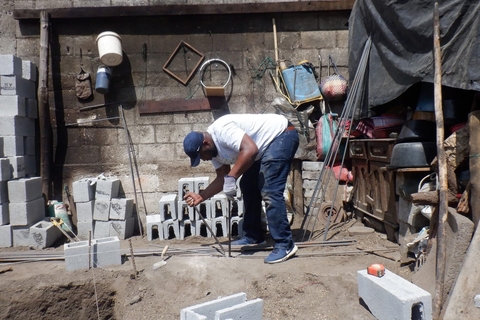 A volunteer bends steel rebar for construction beside stacked concrete blocks and tools.