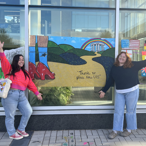 Two women standing next to a painted window with a yellow brick road and the text "there's no place like UIS."