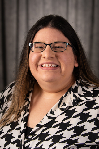 professional head shot of Dr. Maria Duenas; she is smiling, wearing glasses, and has long brown hair and a black and white patterned shirt