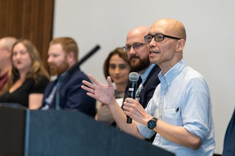 A speaker addresses the audience during a panel discussion, holding a microphone while other panelists listen in the background.