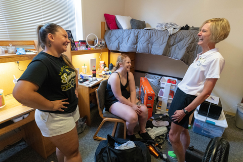 Chancellor Gooch laughs with two female students on move-in day in a residence hall room. Belongings are scattered around the room as they move in for the year.