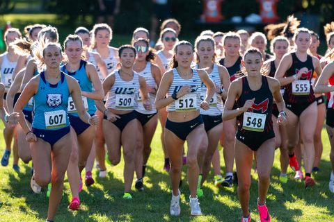 A group of female cross-country runners, including UIS athletes, competes in a race on a sunny day.