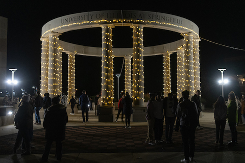 colonnade at night with white lights wrapped around columns and people gathered around it 