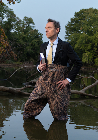 Associate Professor Brandon Derman wears a suit and camouflaged fishing waders while standing in a placid pool of water with trees in the background. 