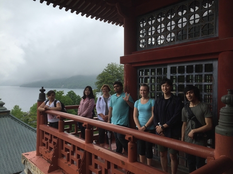 Seven students/staff members standing outside a red Japanese building overlooking a lake on a cloudy day