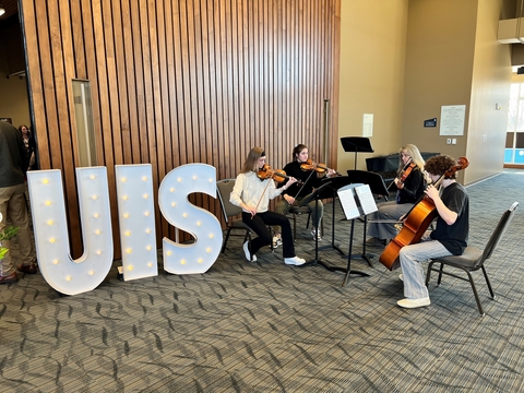 Four students sitting in chairs playing musical instruments. To the left of the students are large letter signs that spell out UIS. 