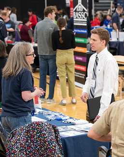 A job fair attendee wearing business attire speaks with a representative at an informational table.