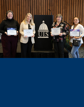 Four award recipients pose with their certificates in front of a UIS podium at a recognition event.