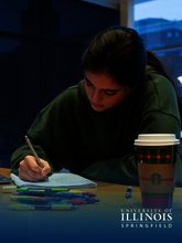 Student studying at a desk with a laptop and coffee cup, in dim lighting.