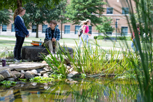 People sitting by a pond with greenery and a building in the background.