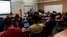 Students in a classroom, some raising hands as a teacher lectures from the front.