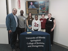 Group photo of MBA students and faculty at the Scoop & Socialize event, standing behind a University of Illinois Springfield College of Business and Management table.
