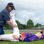 Baseball coach helps player stretch leg on field, cloudy sky.