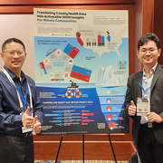 Two men smiling and giving thumbs up, standing beside a colorful conference poster on a stand.