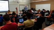 Classroom with a teacher lecturing and students seated, some raising hands.