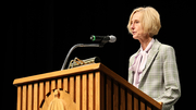 Blonde woman speaking at a wooden podium with a microphone.