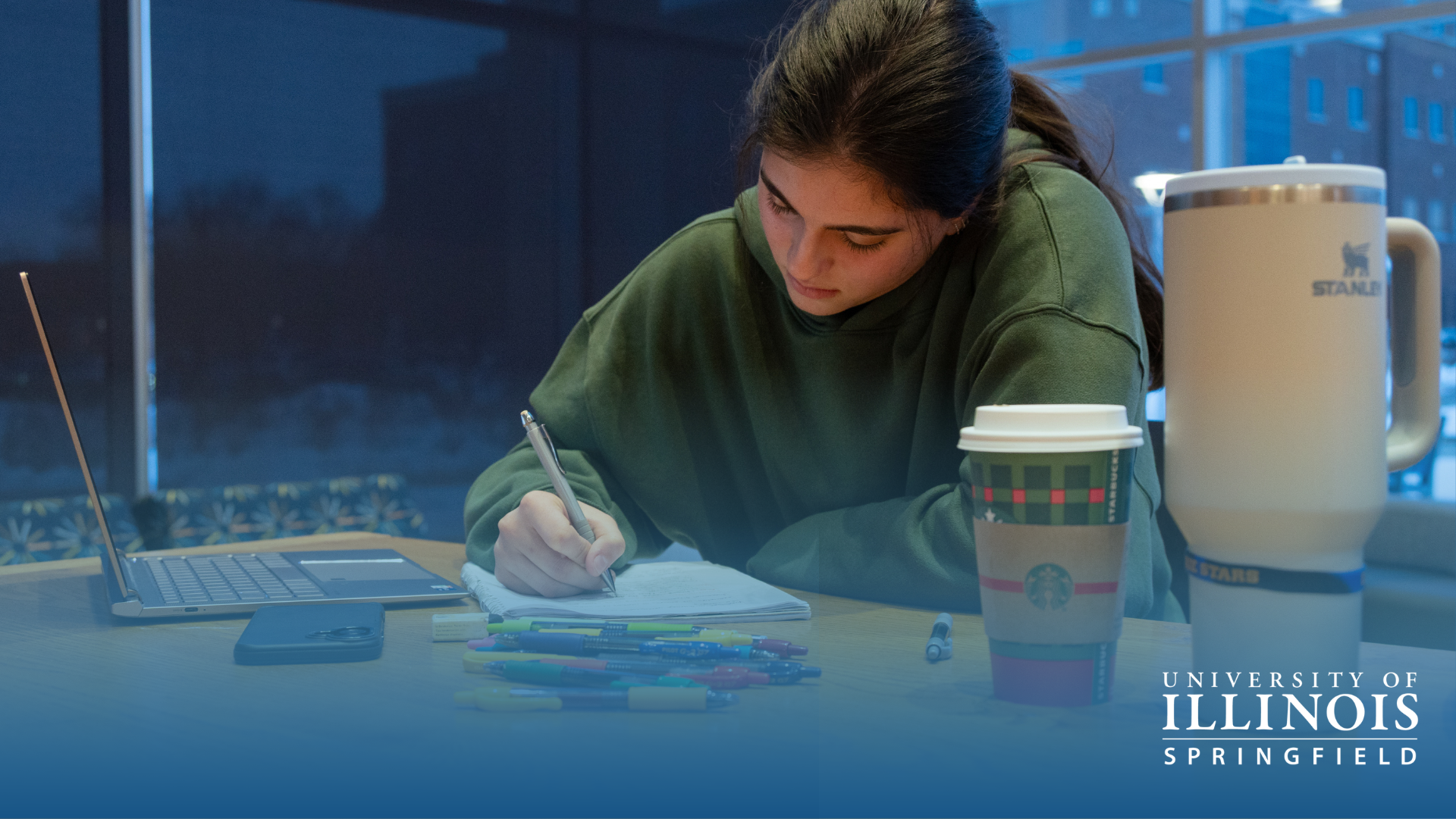 Student studying at a desk with a laptop and coffee cup, in dim lighting.