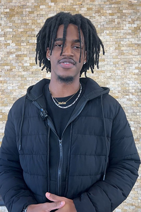 Young man in a black jacket standing against a brick wall.