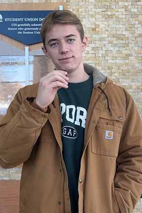 Young man in a brown jacket stands indoors, smiling gently.