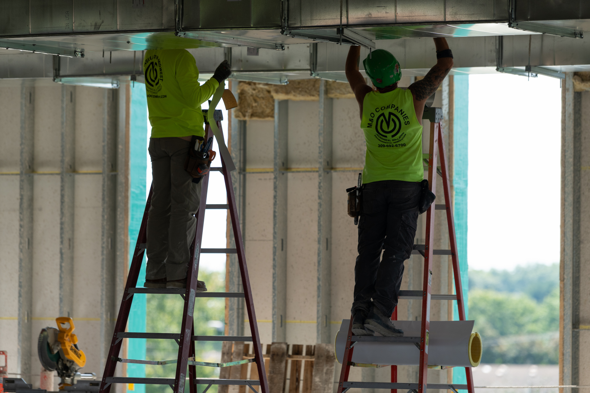 Two construction workers on ladders install overhead ductwork inside the UIS Library Commons.