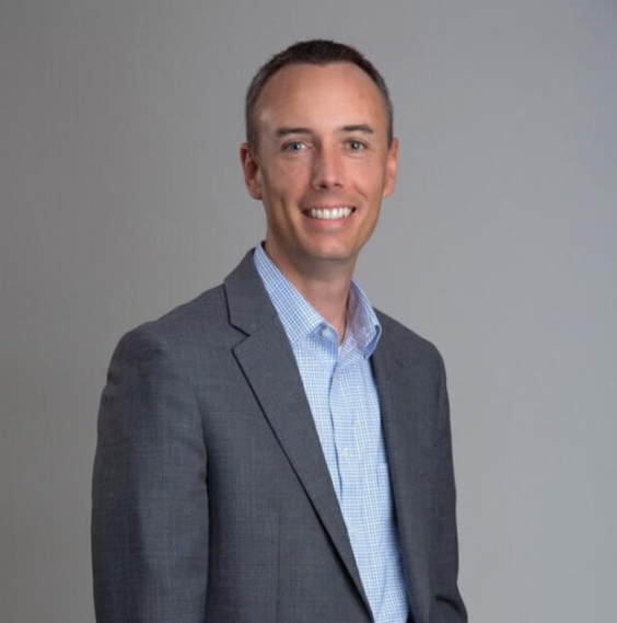Smiling man in a dark suit and blue shirt against a gray background.