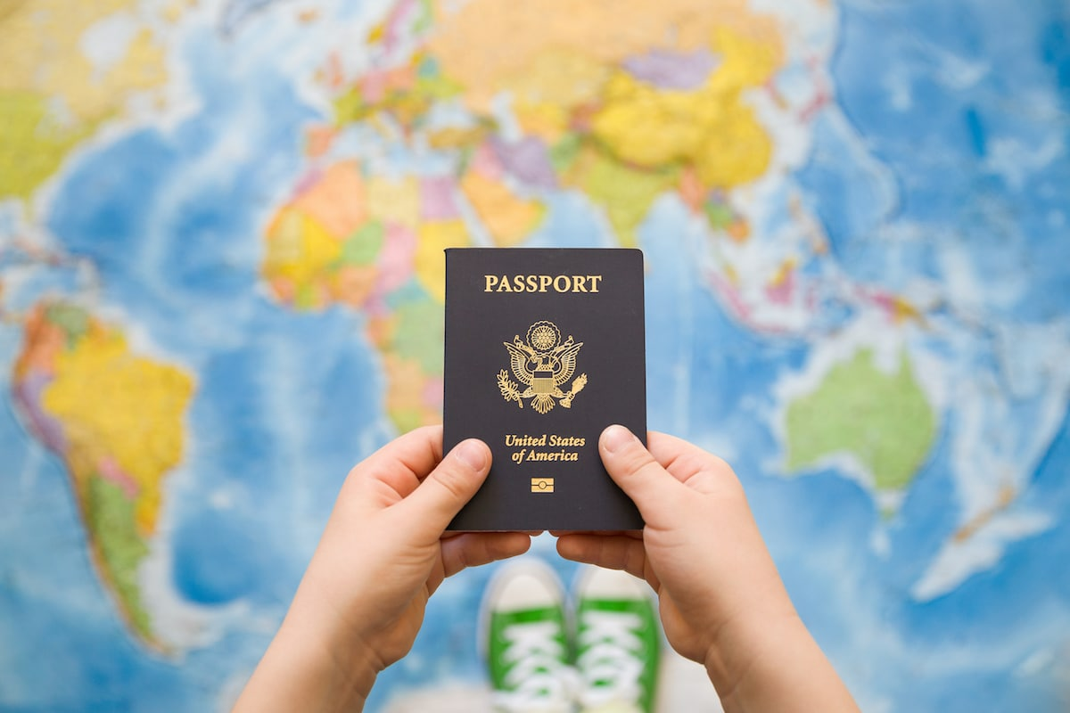 A person holding a US passport in front of a world map. The person is wearing bright green converse shoes.