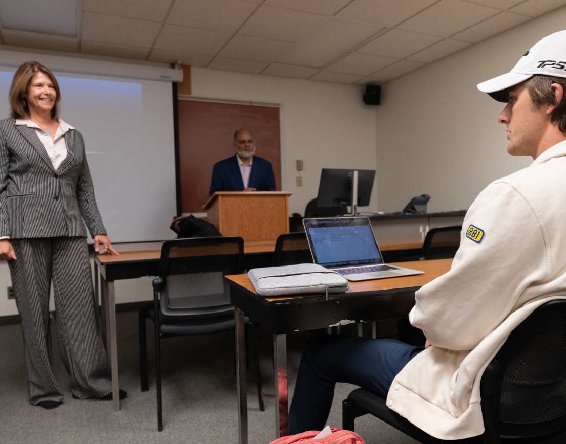 Professor standing at front of class in suit, looking at student sitting in white hoodie and hat. 