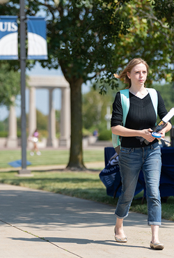 A student carrying books walking on a sidewalk with the colonnade in the background.