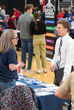 A job fair attendee wearing business attire speaks with a representative at an informational table.