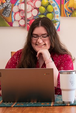 A person in glasses, wearing a red shirt, smiles while working on a laptop at a table with a white travel mug nearby.