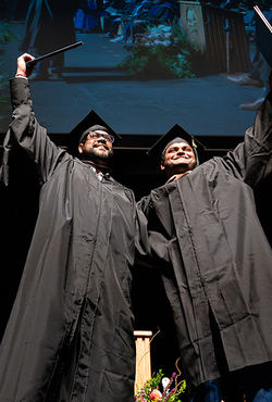 Two graduates in black caps and gowns celebrate on stage by raising their diplomas triumphantly, smiling as they stand side by side. Behind them, faculty members in academic regalia sit in rows and a large screen displays a projection of the moment above them.