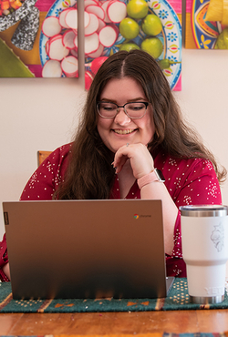 Student smiling while working on a laptop at a table with a tumbler, colorful art hanging on the wall behind her.
