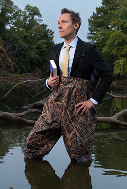 Brandon Derman stands in water wearing waders and a suit, holding a book, with trees in the background.