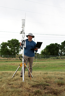 Man with tablet stands by weather station in grassy field.
