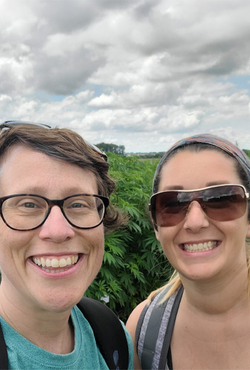 Two people smiling outdoors under a cloudy sky, surrounded by greenery.