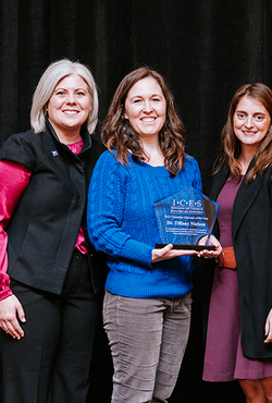 Three women smiling, one holding an award in a blue sweater. Black background.