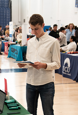Young man reading a brochure at a busy indoor event with booths.