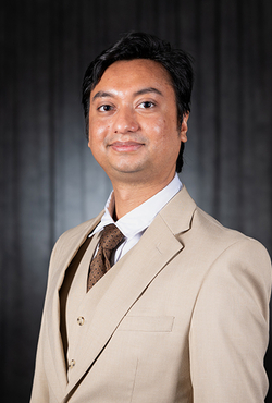 Man in a tan suit with a tie, black background, smiling gently.